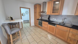 A modern kitchen with wooden cabinets, a black countertop, stainless steel appliances, and a dining area with a bench and table. - Vacation apartment AU (Bitburg)