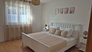 A bedroom with a bed, bedside tables, a nightstand, a lamp, and a window with sheer curtains. - Vacation apartment AU (Bitburg)