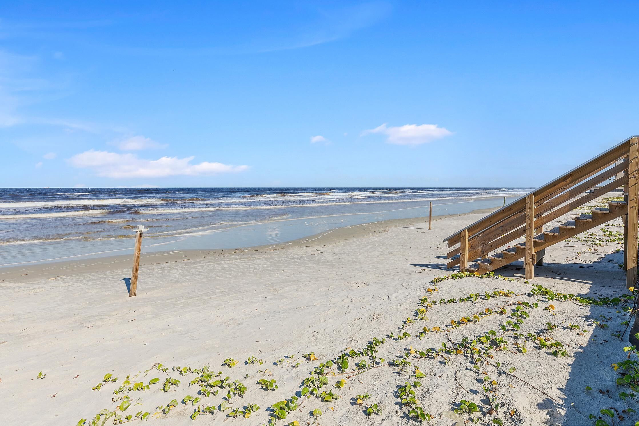 A sandy beach with a wooden staircase leading down to the water, a blue lifeguard stand, and a clear blue sky.