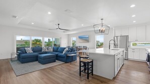 A spacious living area with a large blue sectional sofa, a kitchen with white cabinets, and a view of the outdoors.