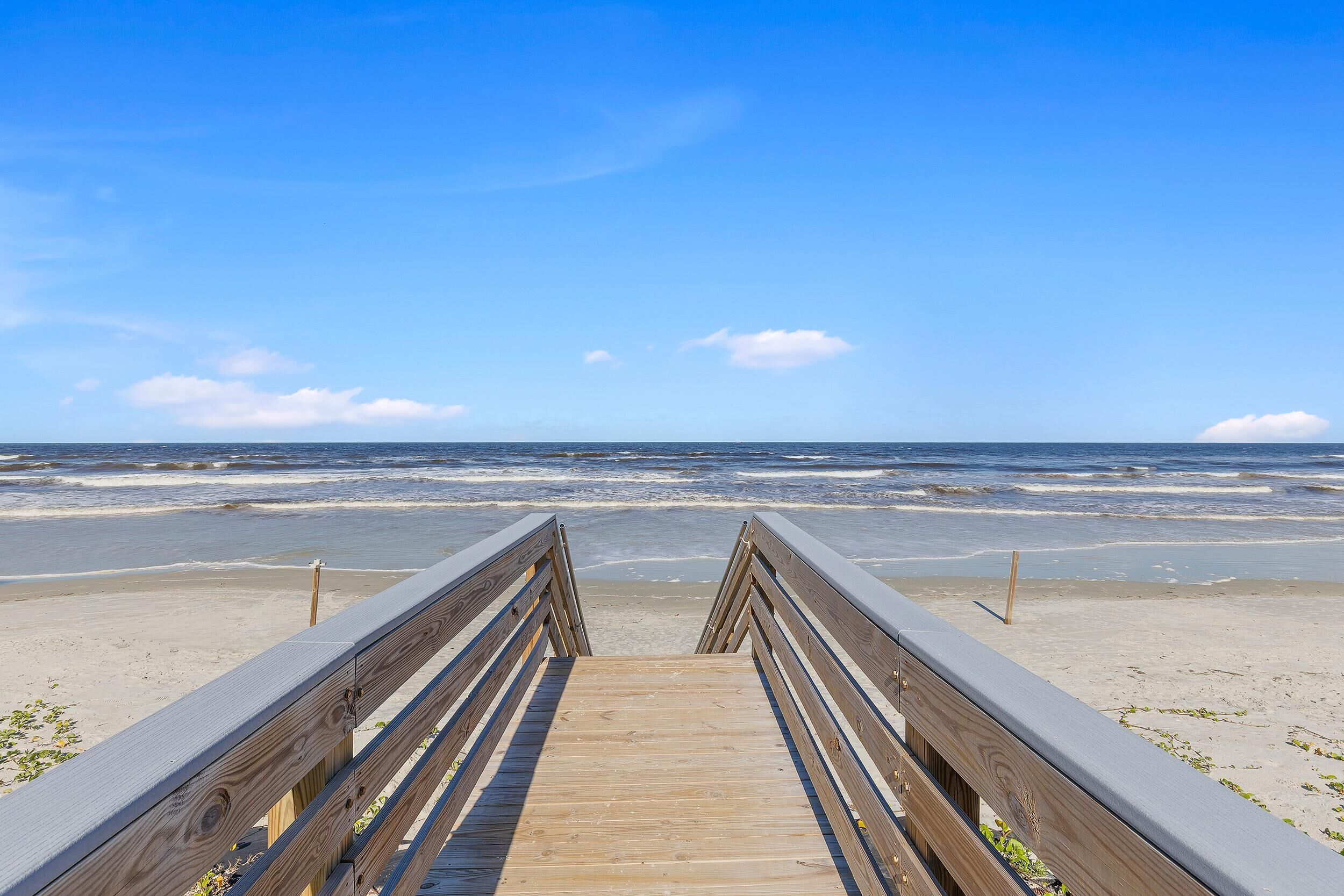 Ein hölzerner Steg führt zu einem Strand, im Hintergrund sind Wellen zu sehen.