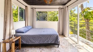 Quarto com cama, mesa de cabeceira, luminária e grandes janelas com vista para o verde.