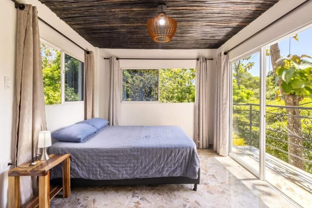 Une chambre à coucher comprenant un lit, une table de chevet, une lampe et de grandes fenêtres donnant sur un paysage verdoyant.