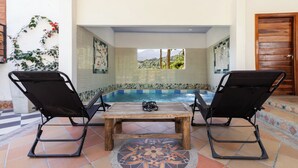 Uma área de piscina com duas espreguiçadeiras pretas, uma mesa de madeira e um chão de azulejos com padrão decorativo.