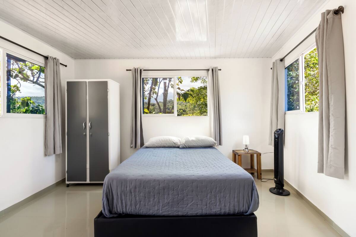 Une chambre à coucher comprenant un lit, une armoire, une table de chevet et un ventilateur sur pied.