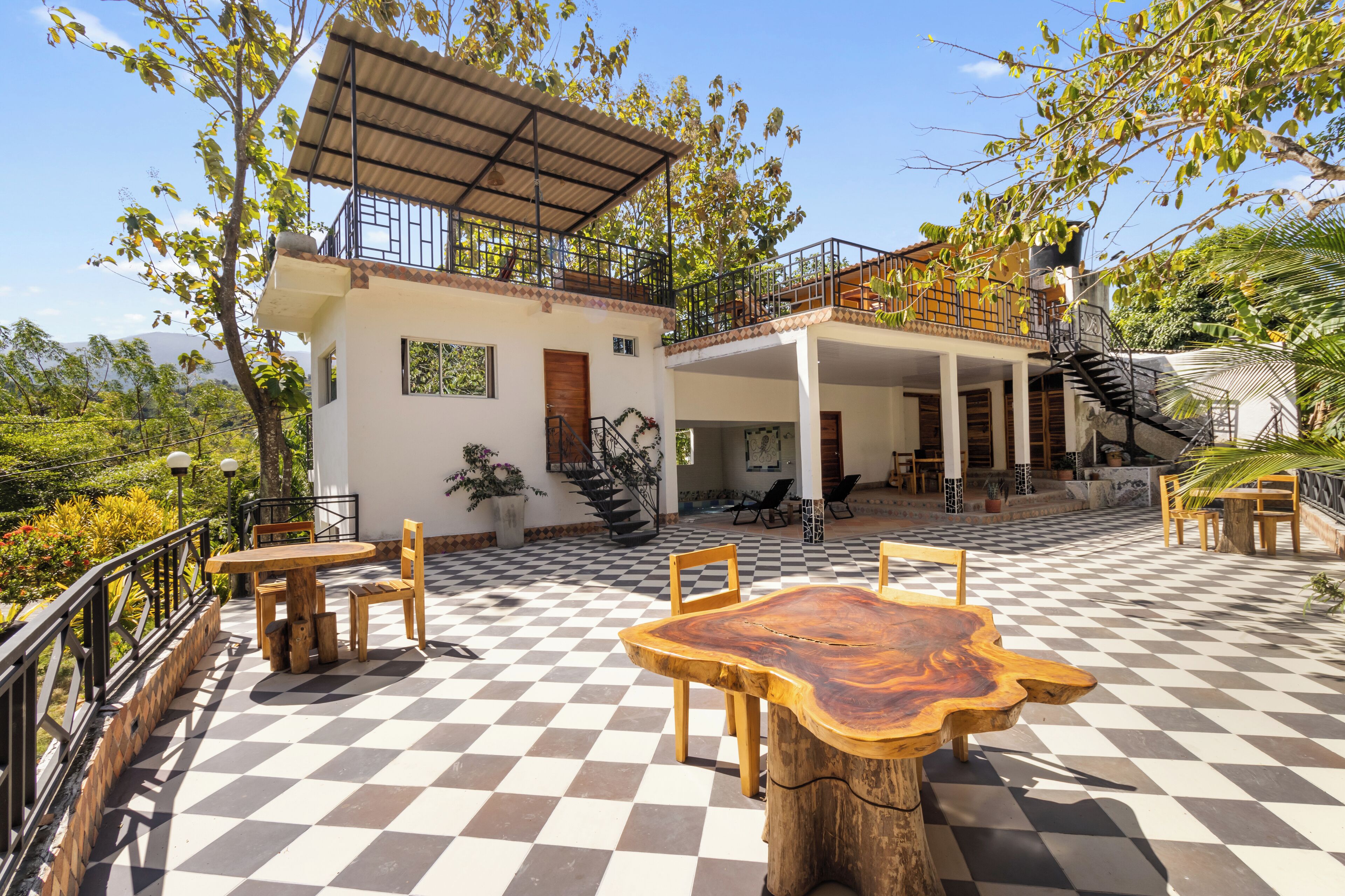 Une vaste terrasse extérieure dotée d’un sol à carreaux, de tables et de chaises en bois, ainsi que d’un espace salon couvert auquel on accède par un escalier menant à un niveau supérieur.