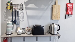 A kitchen with a metal rack holding pots, a wooden cutting board, a toaster, a kettle, and a fire blanket.