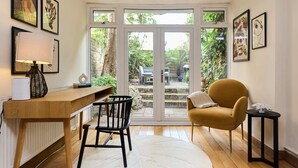 Una habitación con un escritorio de madera, una silla, un sofá y una mesita pequeña. Hay cuadros en las paredes y vistas al exterior a través de un ventanal.
