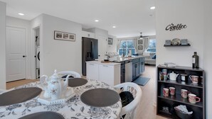 A modern kitchen with white cabinetry, a dining area with a floral tablecloth, and a coffee-themed shelf.