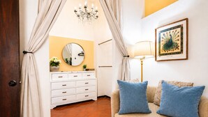 Une pièce avec un canapé beige, deux coussins bleus, une commode blanche à tiroirs, un miroir rond, un lustre et un tableau représentant un paon, encadré, accroché au mur.