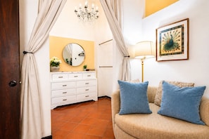 Une pièce avec un canapé beige, deux coussins bleus, une commode blanche à tiroirs, un miroir rond, un lustre et un tableau représentant un paon, encadré, accroché au mur.
