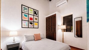 Une chambre à coucher comprenant un lit, une table de chevet, une lampe fixée au mur, une fenêtre donnant sur un paysage agréable et une porte avec un volet en bois.