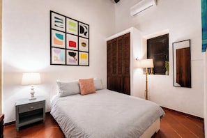 Un dormitorio con cama, una mesita de noche, una lámpara de pared, una ventana con vistas y una puerta con persiana de madera.