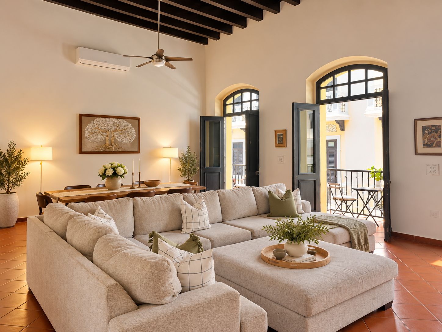 Un salon comprenant un canapé d’angle, une table à manger avec des chaises, un ventilateur de plafond et une vue sur un balcon extérieur accessible par des portes ouvertes.