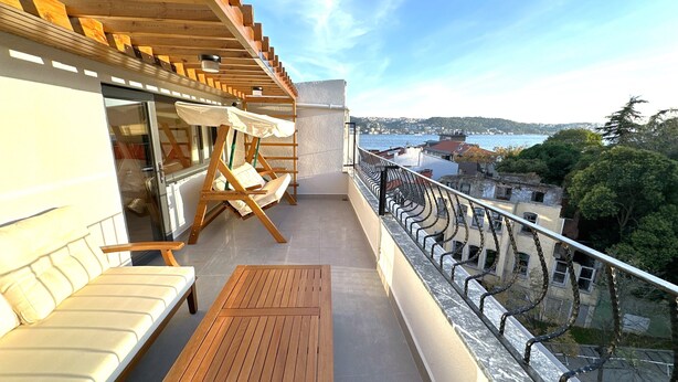 A balcony with a wooden table and chairs, a white sofa, and a view of the sea and buildings. - Flex Ortaköy (İstanbul)