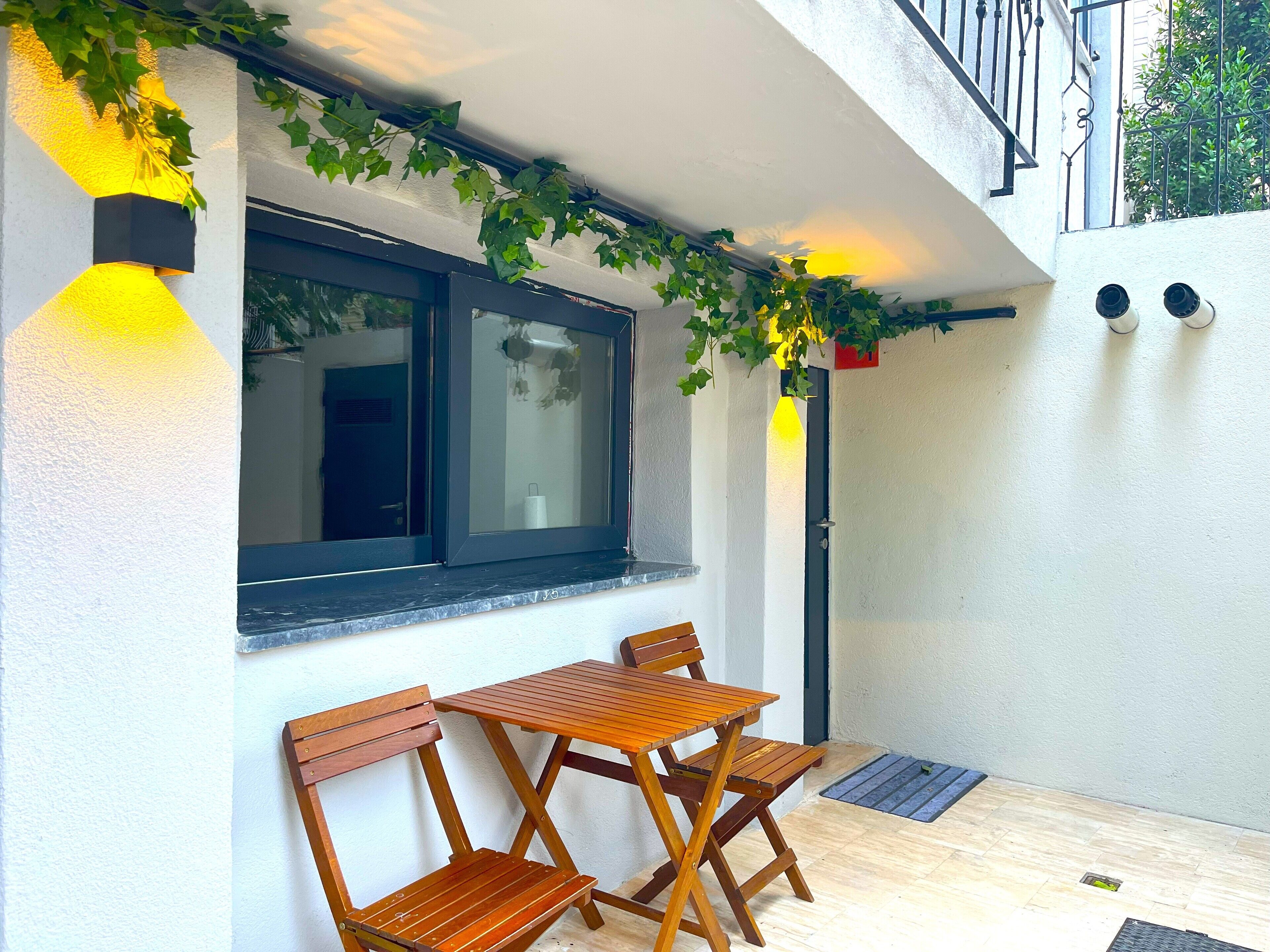 A small outdoor seating area with a wooden table and chairs, a window with a view, and a white wall with a textured surface.