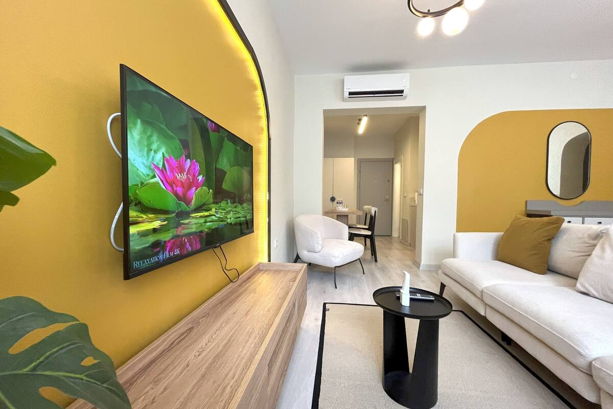 Una sala moderna con un televisor de pantalla plana montado en una pared amarilla, un sofá blanco, una pequeña mesa negra y una zona de comedor al fondo.