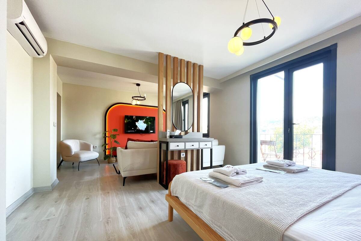 Una habitación de hotel moderna con una cama grande, un escritorio con una silla, un televisor montado en una pared de acento color naranja, y un balcón con barandilla.