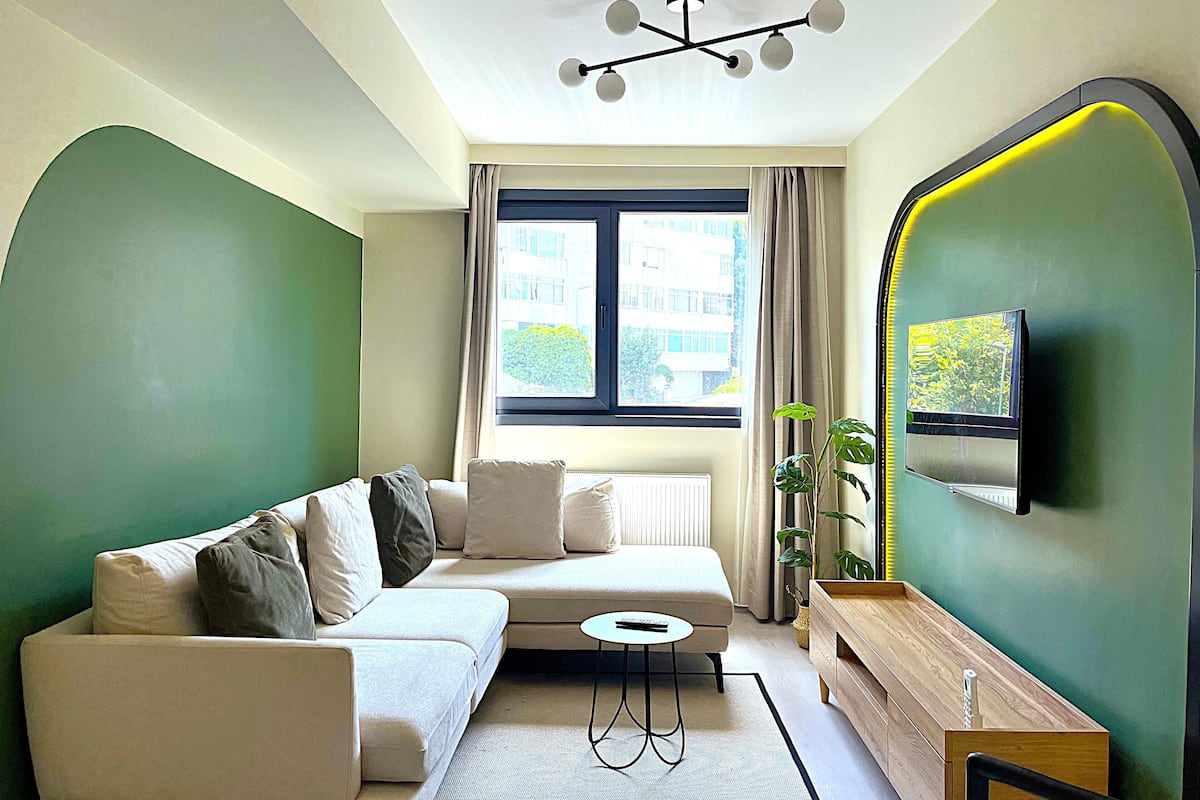 Una sala de estar moderna con un sofá, una mesita, un televisor montado en la pared y una ventana con cortinas.