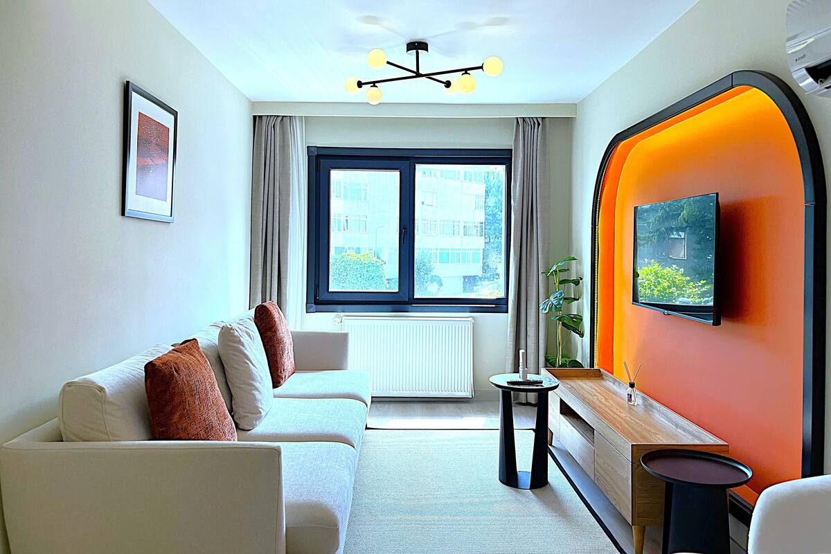 Una sala de estar moderna con un sofá, una mesita auxiliar, un televisor montado y una ventana con cortinas.