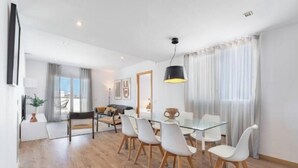 Un salon moderne avec une table à manger en verre, des chaises blanches, un canapé et une grande fenêtre avec des rideaux.
