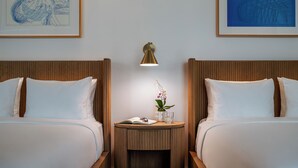 A hotel room with two beds, a nightstand with a lamp, a framed artwork above the bed, and a small table with a plant and a glass of water.