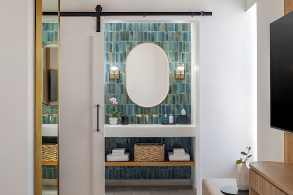 A bathroom with a large mirror, blue-tiled wall, and a shelf with towels and a basket.
