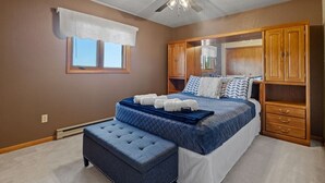 Un dormitorio con cama, cómoda, ventana con cortinilla blanca y ventilador de techo.