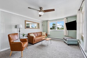Ein Wohnzimmer mit braunem Ledersofa, einem Holztisch, einem braunen Ledersessel, einem an der Wand befestigten Flachbildfernseher und einem Deckenventilator.