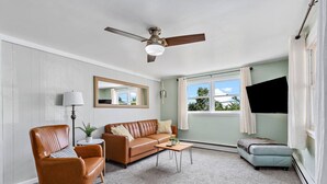 Ein Wohnzimmer mit einem braunen Ledersofa, einem Holztisch, einem braunen Leder-Sessel und einem hellblauen Sofa. Ein Deckenventilator ist sichtbar, und es gibt ein großes Fenster mit Vorhängen.