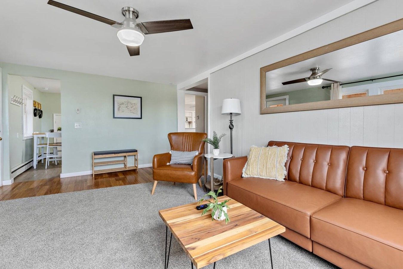 A living room with a brown leather sofa, a wooden coffee table, a brown leather armchair, a ceiling fan, and a large mirror on the wall.