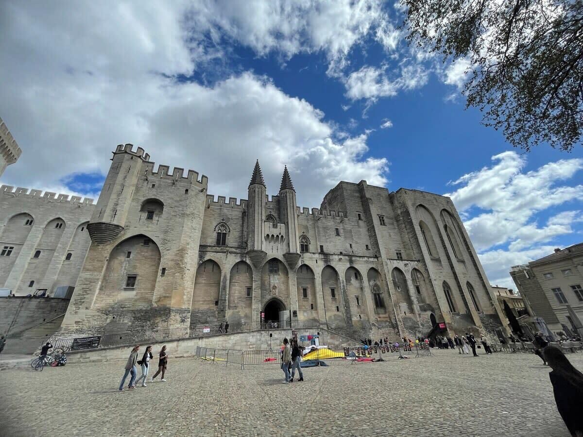Un bâtiment historique en pierre, doté de nombreuses arcades et de tours, qui ressemble probablement à un château ou à une forteresse, avec une cour pavée à l’avant.