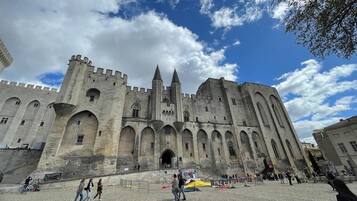 Un bâtiment historique en pierre, doté de nombreuses arcades et de tours, qui ressemble probablement à un château ou à une forteresse, avec une cour pavée à l’avant.