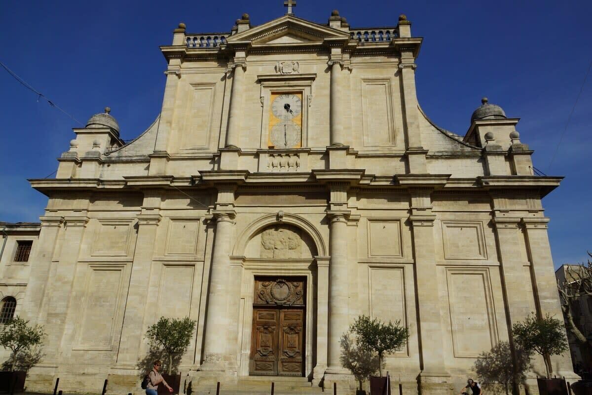 Un edificio grande e histórico con un reloj destacado, fachada simétrica y una entrada central con una puerta ornamentada.
