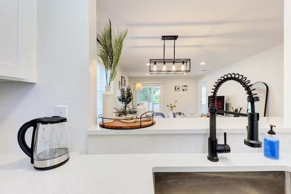 A modern kitchen with a white countertop, a black kettle, a sink with a black faucet, and a hanging light fixture.
