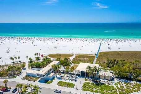 An aerial view of a beach with a large number of people, a clear blue sky, and a calm sea. There are buildings and palm trees visible in the foreground.