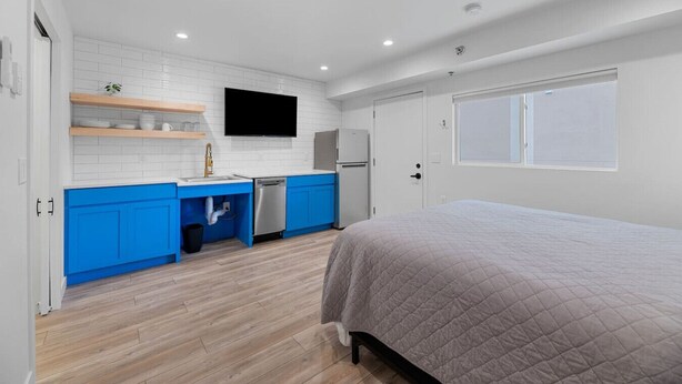 Un dormitorio compacto con cama, una pequeña kitchenette con lavaplatos y refrigerador, y un televisor montado en la pared.
