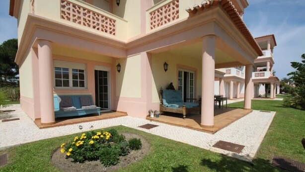 A two-story building with a covered patio area, a small garden with yellow flowers, and a clear sky. - Casa Del Rey - Golf and Beach (Amoreira)