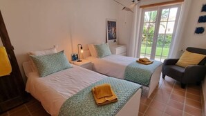 A bedroom with two beds, a nightstand, a chair, and a window with a view of greenery. - Casa Del Rey - Golf and Beach (Amoreira)