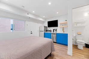 A modern bedroom with a bed, a kitchenette with blue cabinets, a television, and a bathroom with a toilet and sink.