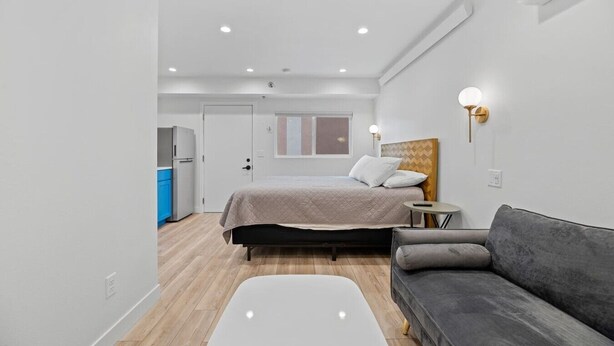 A modern bedroom with a bed, a grey sofa, a small round table, and a white bedside table. - Queen City Inn and Extended Stay #2 (Spearfish)