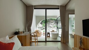 Una moderna camera d'albergo con un letto, una scrivania, una sedia, un'ampia finestra e vista su una zona esterna con piscina e rocce.