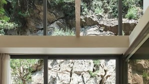 Ambiente moderno con vista su terreno roccioso attraverso ampie finestre, una panca in legno e un tavolino con un libro e una ciotola.