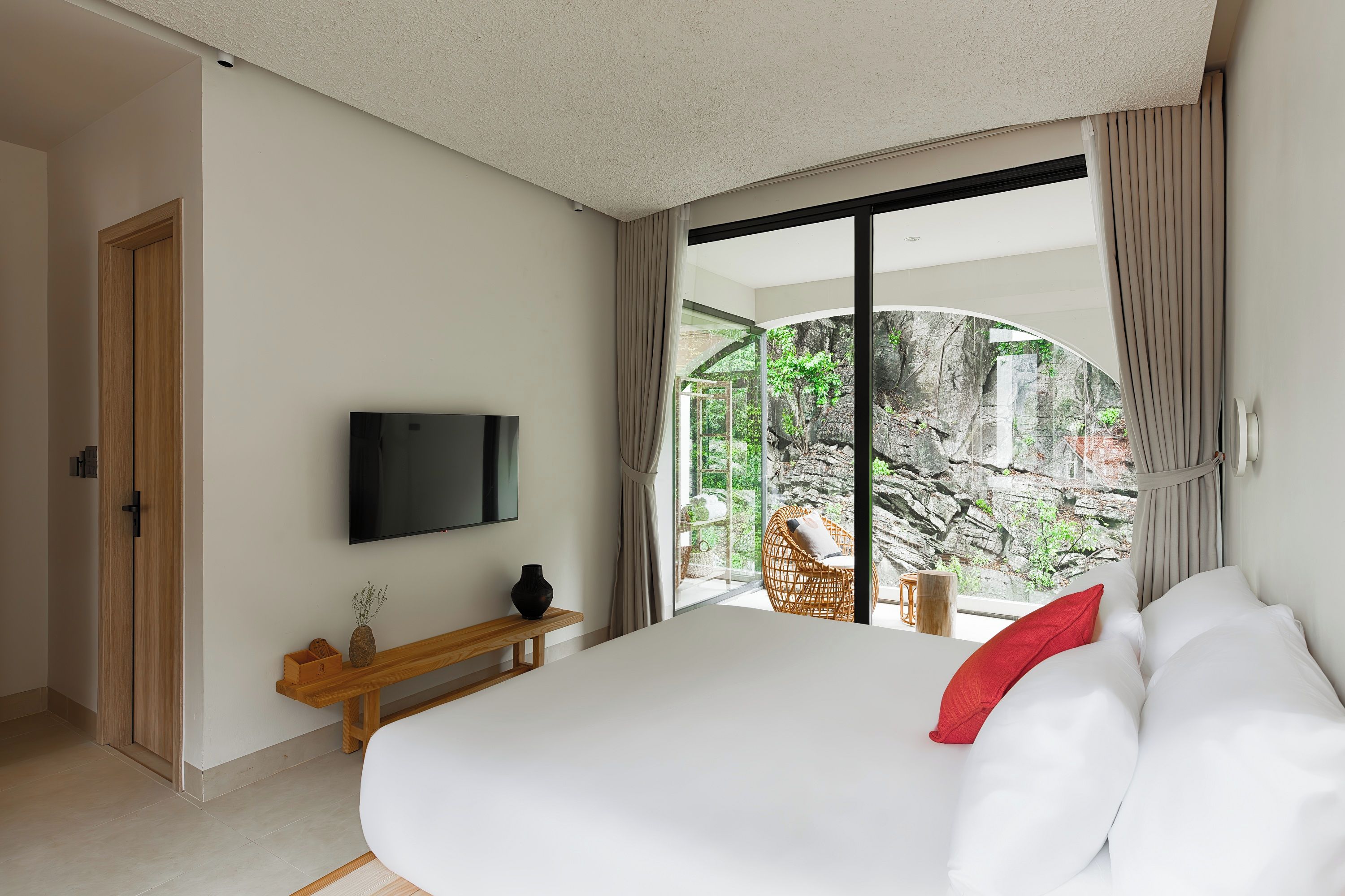 A bedroom with a large bed, a television mounted on the wall, and a view of a rocky outdoor area through a glass door.