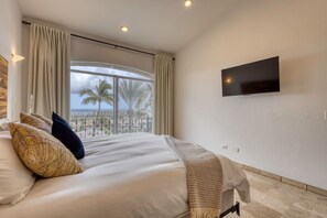A hotel room with a bed, a TV mounted on the wall, and a view of palm trees and the sea.