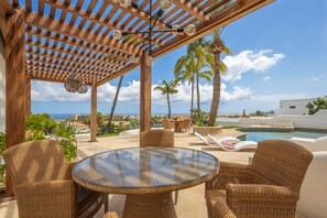 A covered outdoor seating area with wicker furniture, a glass table, and a view of the ocean.