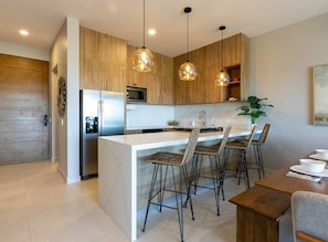 A modern kitchen with a central island, bar stools, pendant lighting, and a dining area with a wooden table. - Modern 2BR with Ocean Views (Cabo San Lucas)