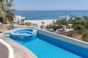 A swimming pool with a view of the ocean and beach, surrounded by palm trees and white buildings.