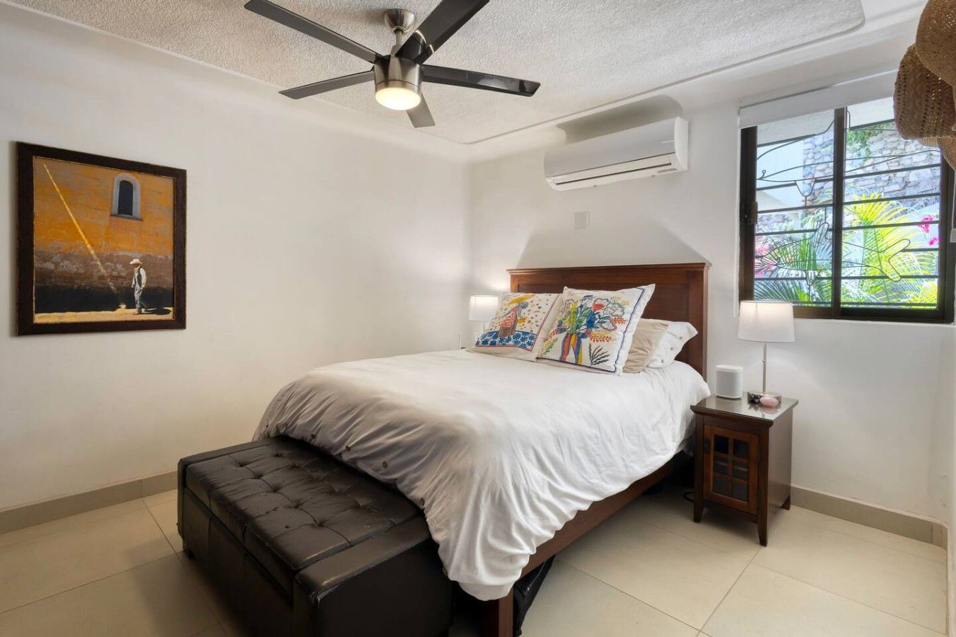 Quarto com cama, ventilador de teto, ar condicionado, janela com vista e um quadro na parede.