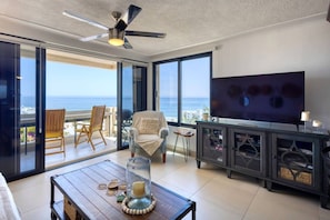 A spacious living room with a large TV, a ceiling fan, and a view of the ocean through large glass doors. - Surfside Azul - Ocean Views (San José del Cabo)